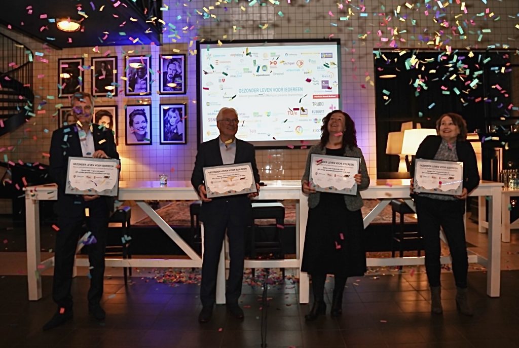 Vier mensen binnen met grote certificaten en confetti. Op de achtergrond een scherm met logo's en tekst, en portretten aan de muur.