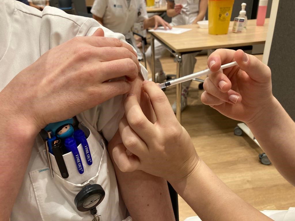 Iemand in wit uniform krijgt een eerste boosterprik in de bovenarm. Pennen en badge aan borstzak, persoon op achtergrond aan tafel.