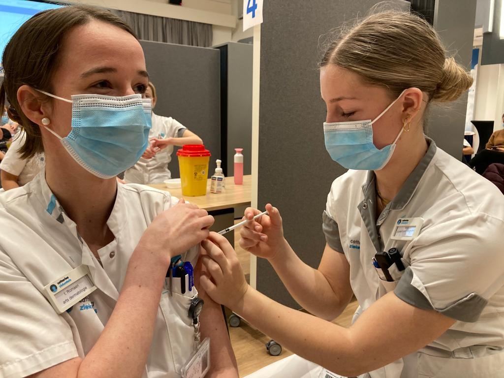 Twee zorgmedewerkers met mondmaskers; een geeft de ander een eerste boostervaccinatie. Medische spullen op tafel.