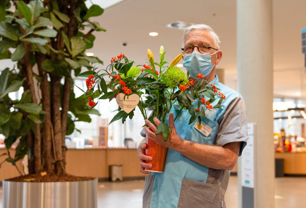 Een oudere man met bril, mondkapje en blauw uniform houdt binnen een bloempot met bloemen vast. Grote plant op achtergrond.