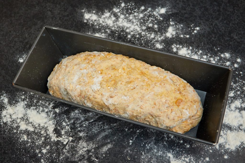 Ongebakken volkoren brooddeeg in een rechthoekige metalen bakblik op een donkere ondergrond met wat bloem.