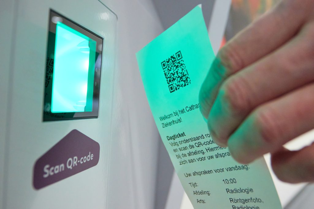 Persoon houdt papier met QR-code bij aanmeldzuil "Scan QR-code" met groen licht; papier toont aanmeld- en afspraakinfo.