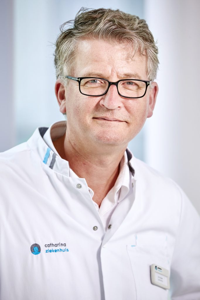 Een man van middelbare leeftijd met licht haar en bril draagt een witte labjas en staat voor een vage achtergrond.