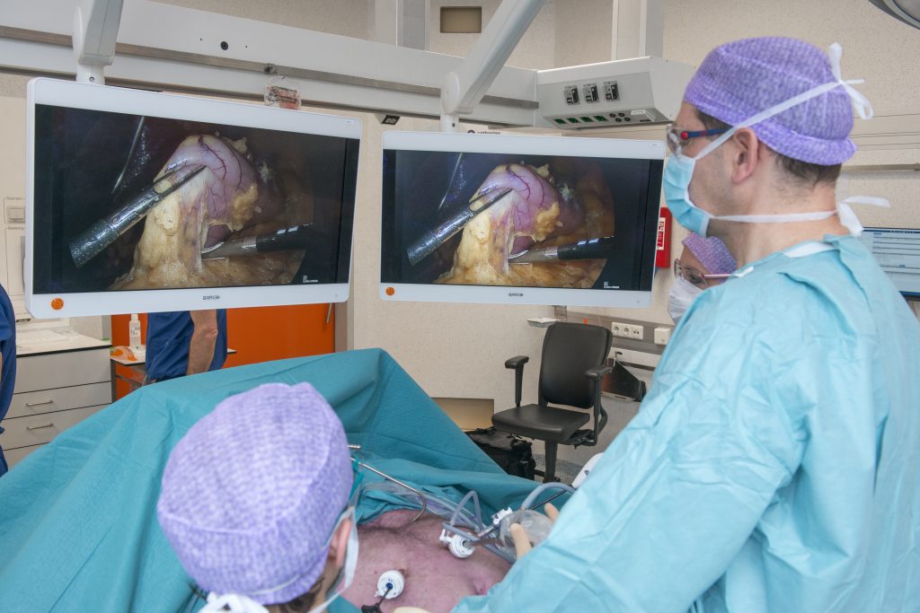 Twee chirurgen voeren een minimaal invasieve operatie uit in een ziekenhuis, met monitors en medische apparatuur op de achtergrond.