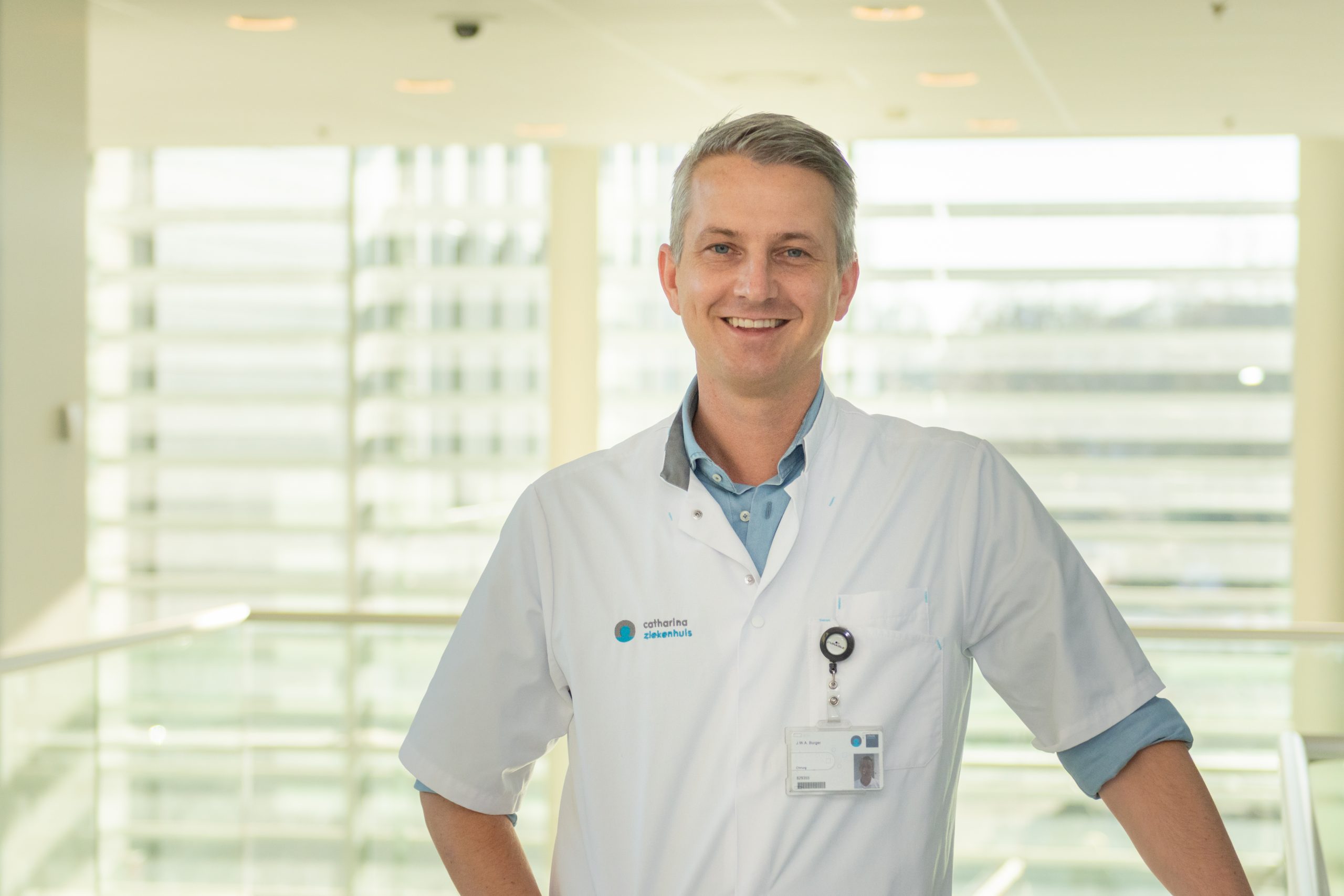 Man in witte doktersjas en ID-badge staat binnen, hand op heup, lachend. Grote ramen laten daglicht binnen.