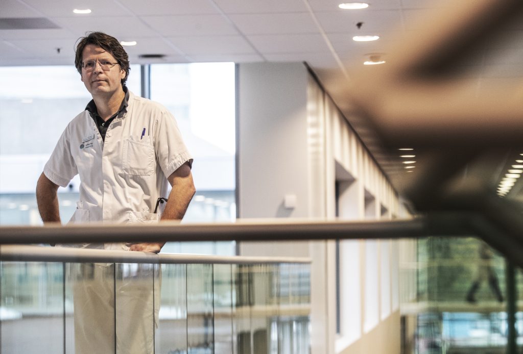 Een persoon in een wit medisch uniform staat bij een reling in een modern gebouw met glazen panelen en veel licht.