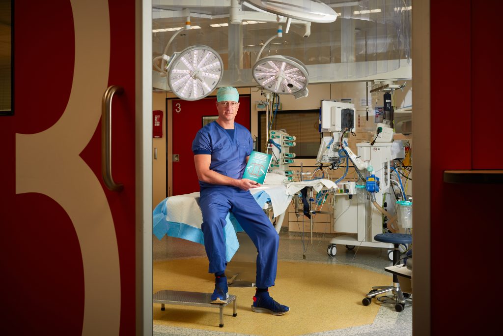 Een hartchirurg in blauwe operatiekleding zit op een kruk in de OK met een boek. Medische apparatuur en rode deuren op de voorgrond.