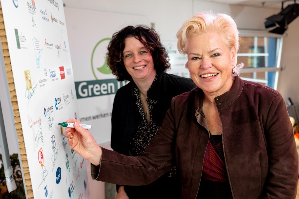 Twee vrouwen staan binnen bij een bord met logo's; één vrouw tekent, beiden lachen. "Groen' en Catharina Ziekenhuis zichtbaar.