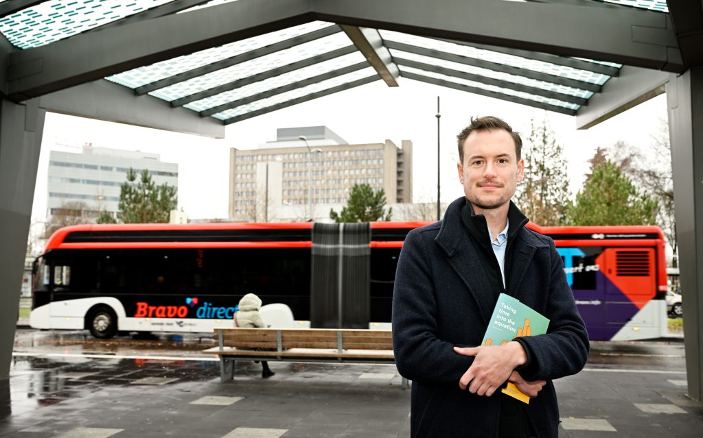 Man in donkere jas onder glazen overkapping, met boek. Op achtergrond rood-zwarte Bravo bus, stadsgebouwen en bomen.