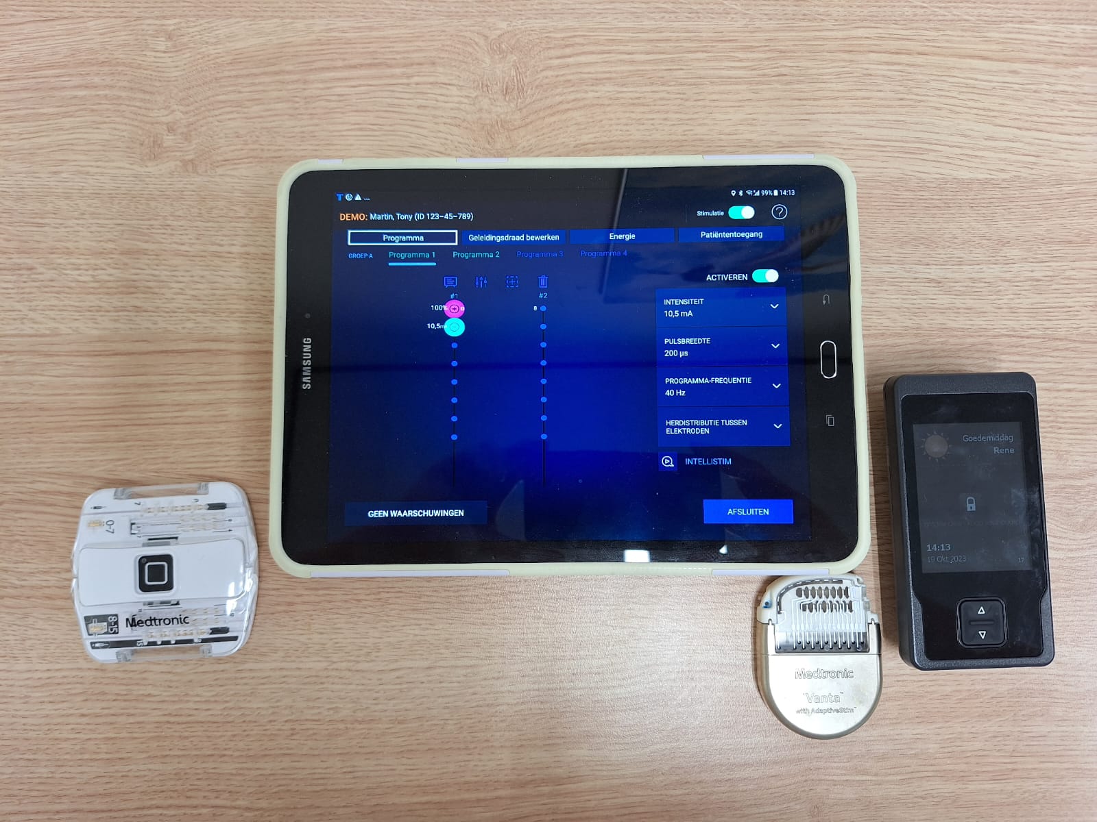 Tablet met medische app op houten ondergrond, omringd door drie neurostimulatoren: wit, zilver ovaal en zwart rechthoekig.