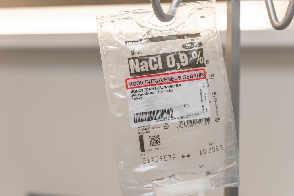 Close-up van een infuuszak 250 ml NaCl 0,9 met Nederlandse labelinformatie en bijwerkingen, hangend aan metalen standaard.