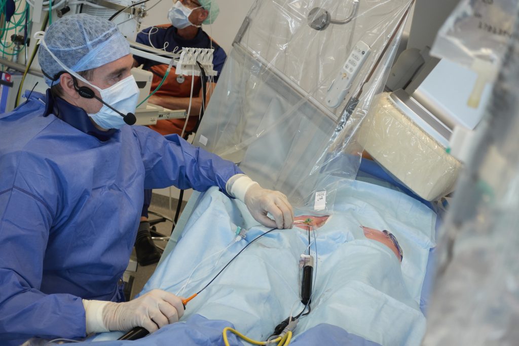 Een zorgverlener in schort en mondkapje voert een procedure uit naast een patiënt; medisch materiaal op de achtergrond zichtbaar.