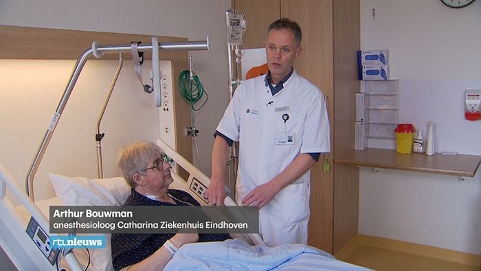 Een arts in witte jas spreekt met oudere man aan ziekenhuisbed. Medische apparatuur op de achtergrond. Namen in beeld.