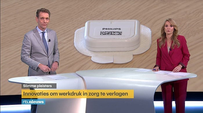 Twee nieuwslezers bij een studiodesk met achter hen een afbeelding van een Philips slimme pleister en Nederlands onderschrift over zorginnovatie.