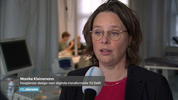Vrouw met bril en bruin haar spreekt in microfoon over slimme pleister; onderschrift: Maaike Kleinsmann, TU Delft. Achtergrond vaag.