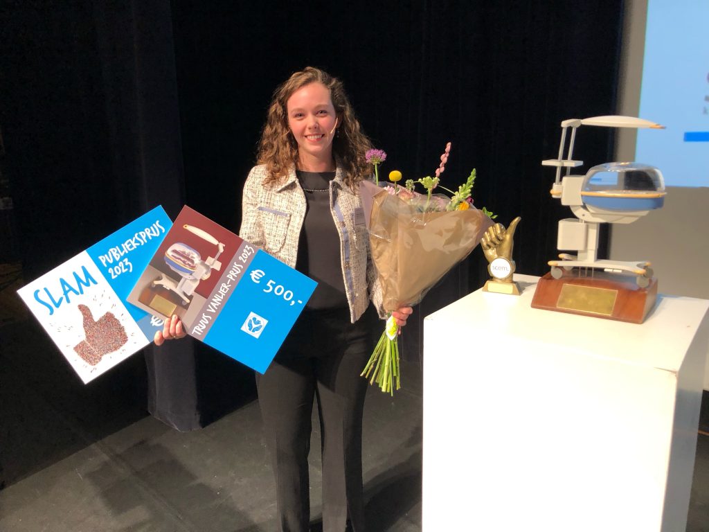 Vrouw in zakelijke kleding op podium met bloemen en cheque, viert SLAM Publieksprijs 2023 bij Catharina Ziekenhuis.