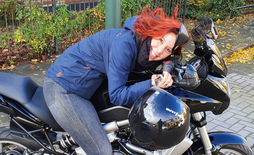 Persoon met rood haar en blauwe jas leunt over zwarte motor en glimlacht. Zwarte helm op tank, herfstbladeren en hek op achtergrond.