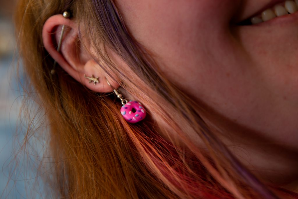 Close-up van een oor met twee oorbellen: een stervormige tap en een roze hanger met paarse stippen. Rood haar met paarse lokken.