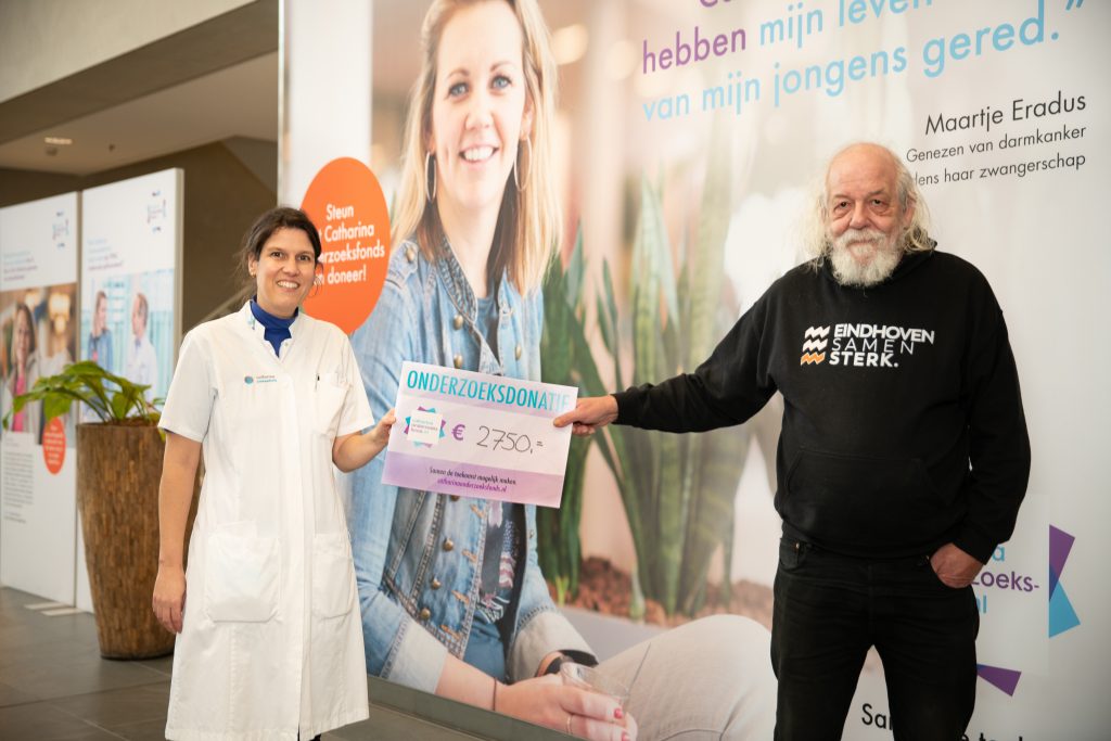 Wonderwoman Naomi in een witte jas en man in zwarte hoodie houden cheque van €2.750 voor Catharina Onderzoeksfonds vast.