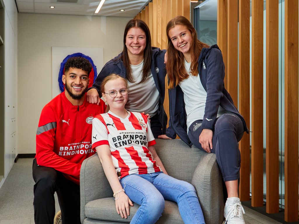 Vier jonge volwassenen binnen; een man en vrouw zitten, twee vrouwen staan achter hen. Allen dragen PSV-shirts.