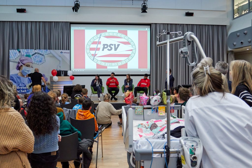PSV-spelers en anderen zitten op een podium voor het PSV-logo, met publiek van medisch personeel en kinderen in het Catharina Ziekenhuis.