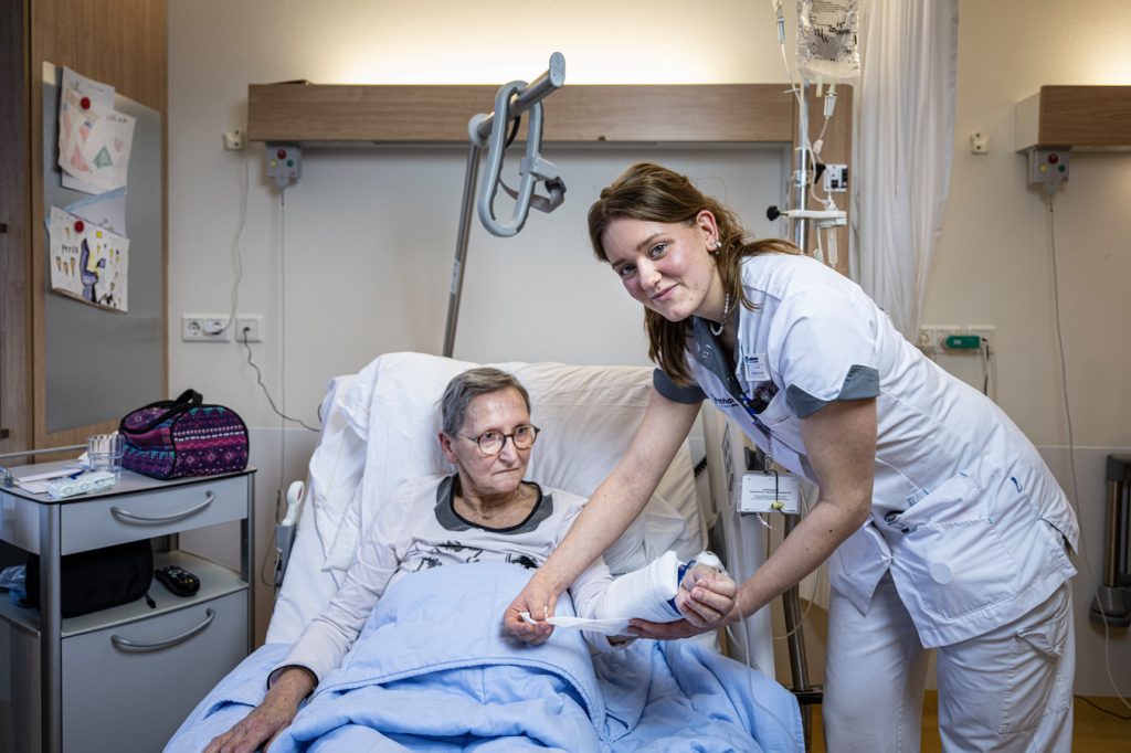 Een verpleegkundige in wit uniform overhandigt papieren aan een oudere vrouw in bed; medische apparatuur en persoonlijke spullen zichtbaar.