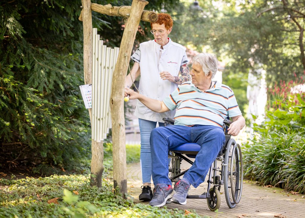 Oudere man in rolstoel slaat met hamer op buitenklokken; zorgmedewerker in wit uniform helpt hem in een groene tuin.