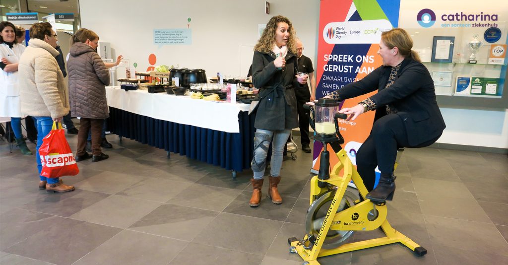 Vrouw in zakelijke kleding op gele smoothiefiets, andere vrouw gebaart. Op achtergrond mensen bij buffet, event in medisch centrum.