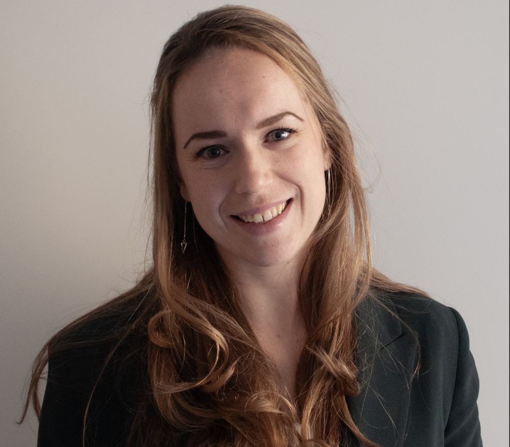 Vrouw met lang, goudbruin haar glimlacht naar de camera. Ze draagt een donkere blazer en oorbellen, neutrale achtergrond.