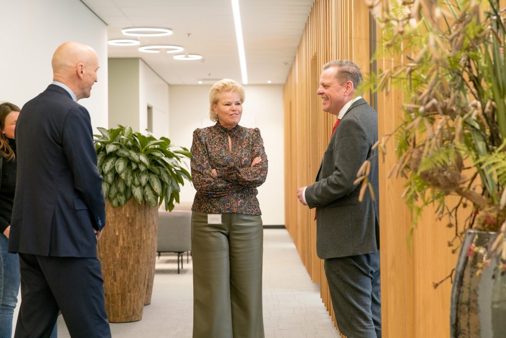 Drie mensen praten in een moderne gang van het Catharina Ziekenhuis tijdens een werkbezoek; planten en hout decoreren de ruimte.