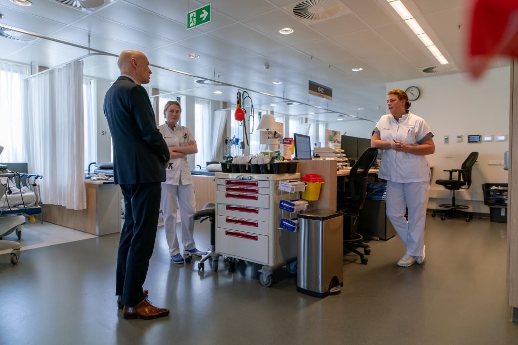 Ernst Kuipers spreekt met twee zorgmedewerkers bij een medicijnkar in een helder verlichte ziekenhuiszaal. Bedden op de achtergrond.