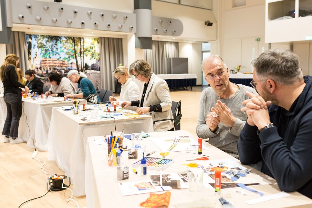Mensen werken creatief aan tafels in een lichte ruimte van het Rijksmuseum; twee mannen met darmkanker praten samen.
