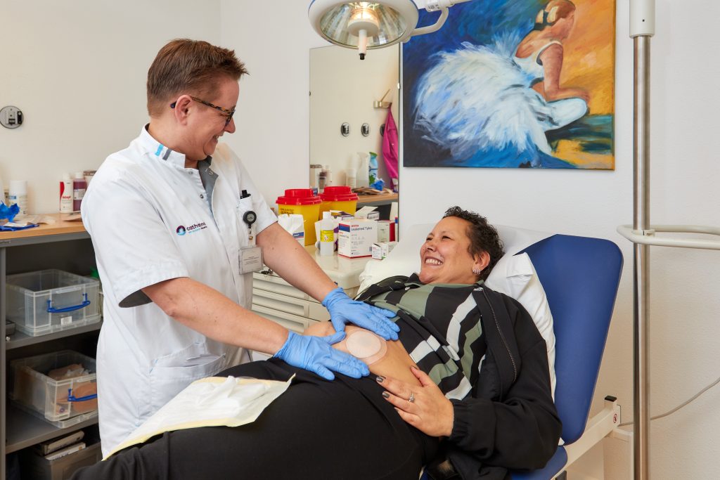 Een zorgverlener met handschoenen verzorgt het verband van een patiënt met stoma, in een medische kamer met schilderij.