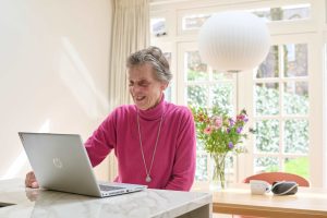 Oudere vrouw in roze trui zit glimlachend aan marmeren tafel achter HP-laptop. Op achtergrond bloemen, lamp en ramen.