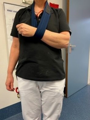 Persoon met zwart shirt en witte broek staat binnen, rechterarm in blauwe mitella. Gesloten deur en medisch affiche op achtergrond.