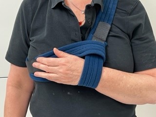 Persoon in zwart shirt met linkerarm in blauwe draagdoek vanwege bovenarmbreuk. Hand onder het sleutelbeen. Lichte achtergrond.