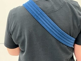 Persoon met zwart shirt van achteren, drie blauwe banden schuin over rechter schouder en rug ter ondersteuning bovenarmbreuk.