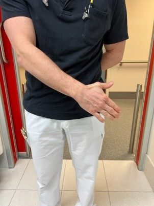 Persoon staat binnen in zwart shirt en witte broek, steekt rechterhand uit voor handdruk. Rode deurkozijnen, beige muren.