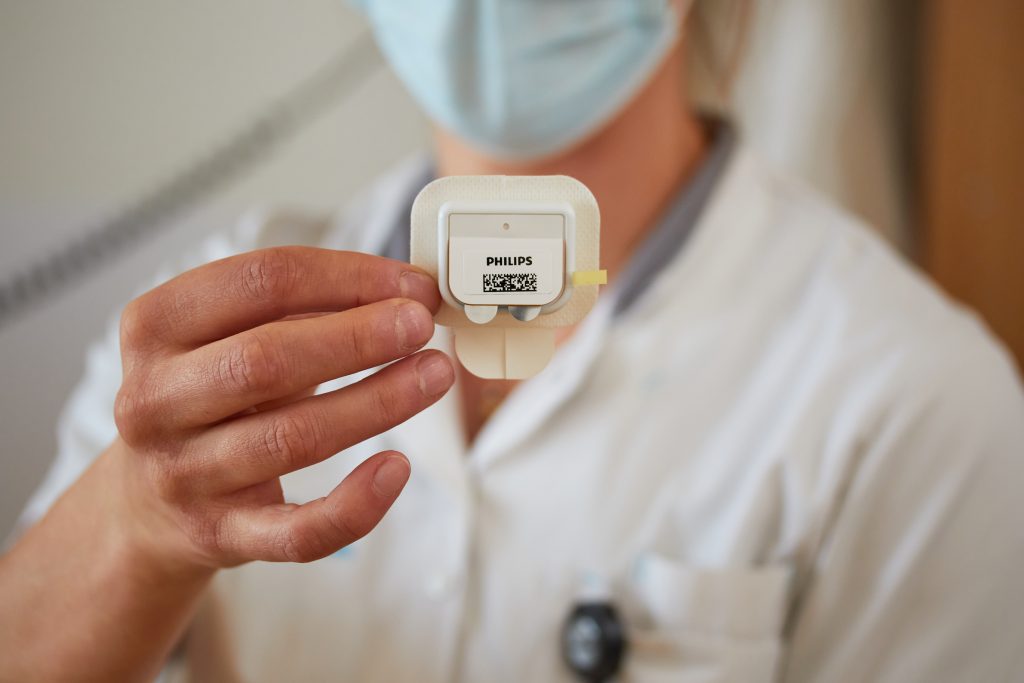 Persoon in witte labjas en mondmasker toont kleine PHILIPS sensor met barcode en QR-code, gebruikt bij patiëntmonitoring.