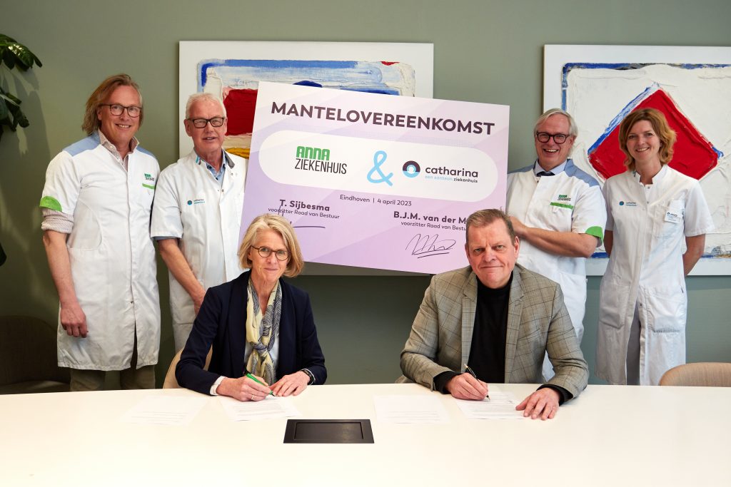 Zes mensen binnen, twee ondertekenen documenten aan tafel, vier staan achter hen. Bord toont logo's en samenwerkingsovereenkomst.