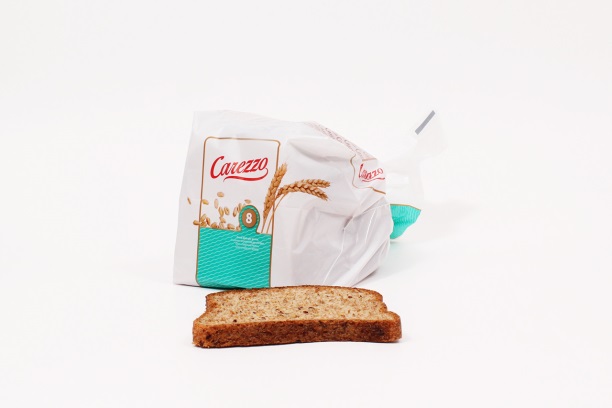 Een sneetje volkorenbrood voor een deels open witte en turquoise Carezzo-broodzak met tarwegrafiek en kaartje.