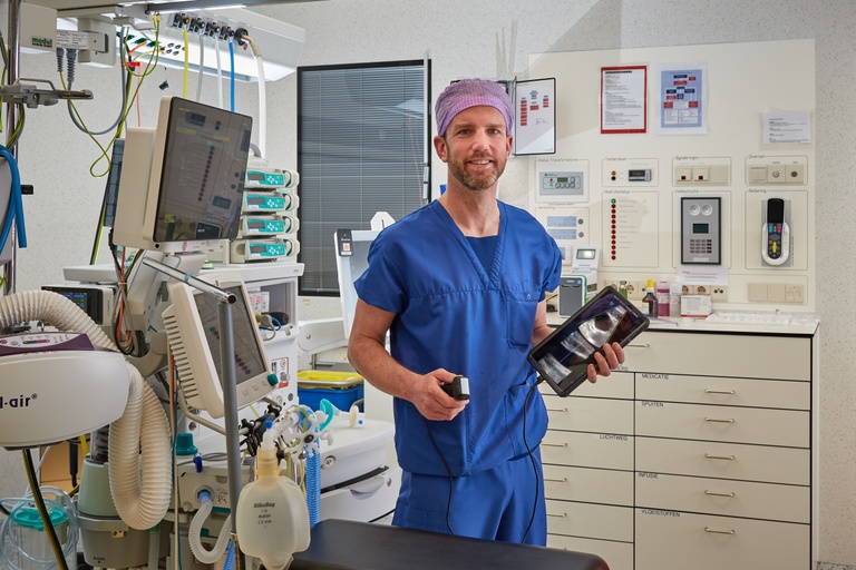 Persoon in blauwe operatiekleding en paarse muts bereidt infuusprik met echografie voor in een behandelkamer.