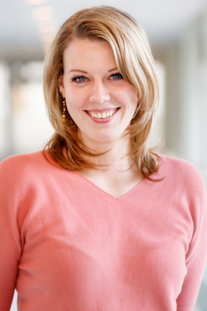 Vrouw met blonde haar en blauwe ogen glimlacht naar de camera. Ze draagt een roze trui, gouden oorbellen en steunt eierstokkanker.
