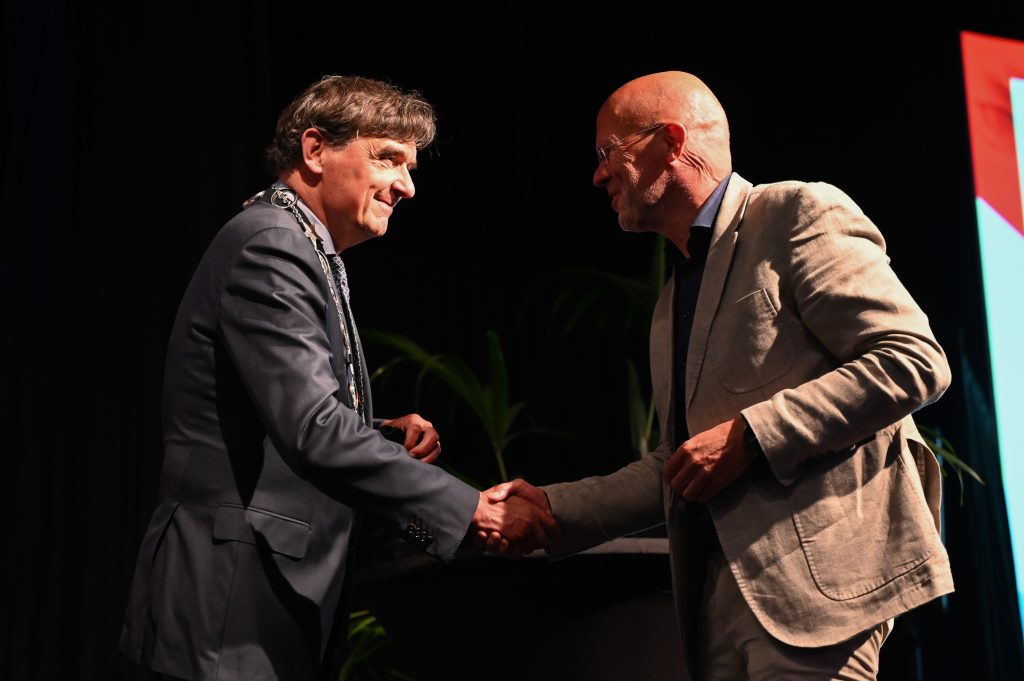 Prof. dr. Joep Teijink met ambtsketen schudt op podium handen bij uitreiking Gouden legpenning, donkere achtergrond.
