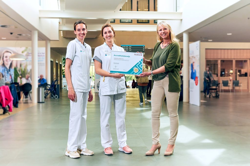 Drie vrouwen in de lobby van het Catharina Ziekenhuis, twee in witte uniformen en één casual, houden lachend een accreditatiecertificaat vast.
