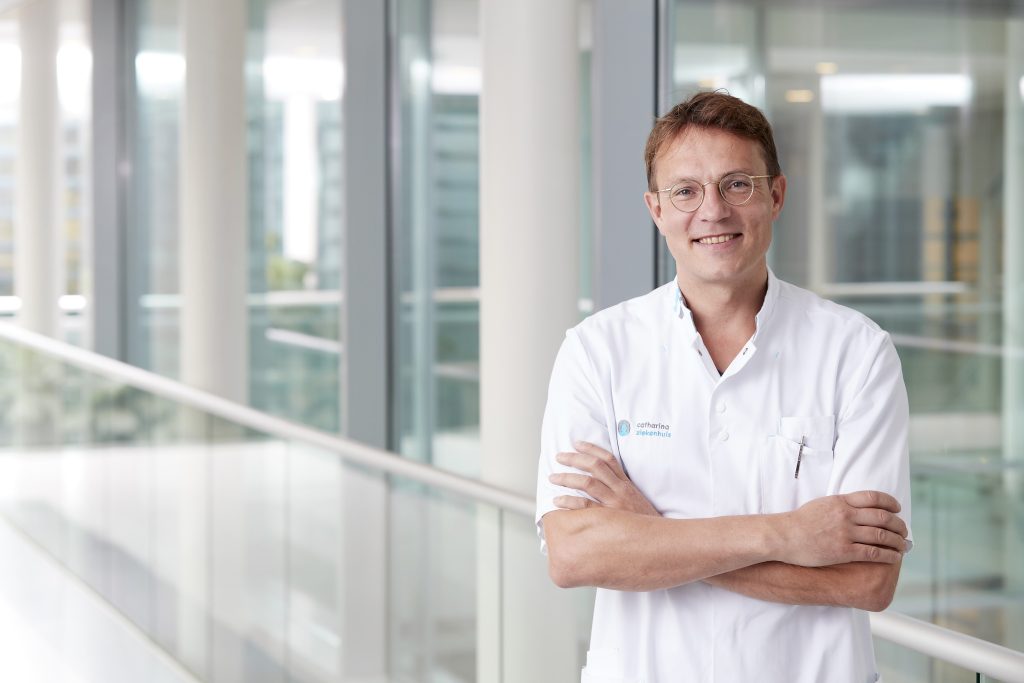 Man met bril en witte doktersjas staat binnen met gekruiste armen in modern gebouw met grote ramen en witte pilaren.
