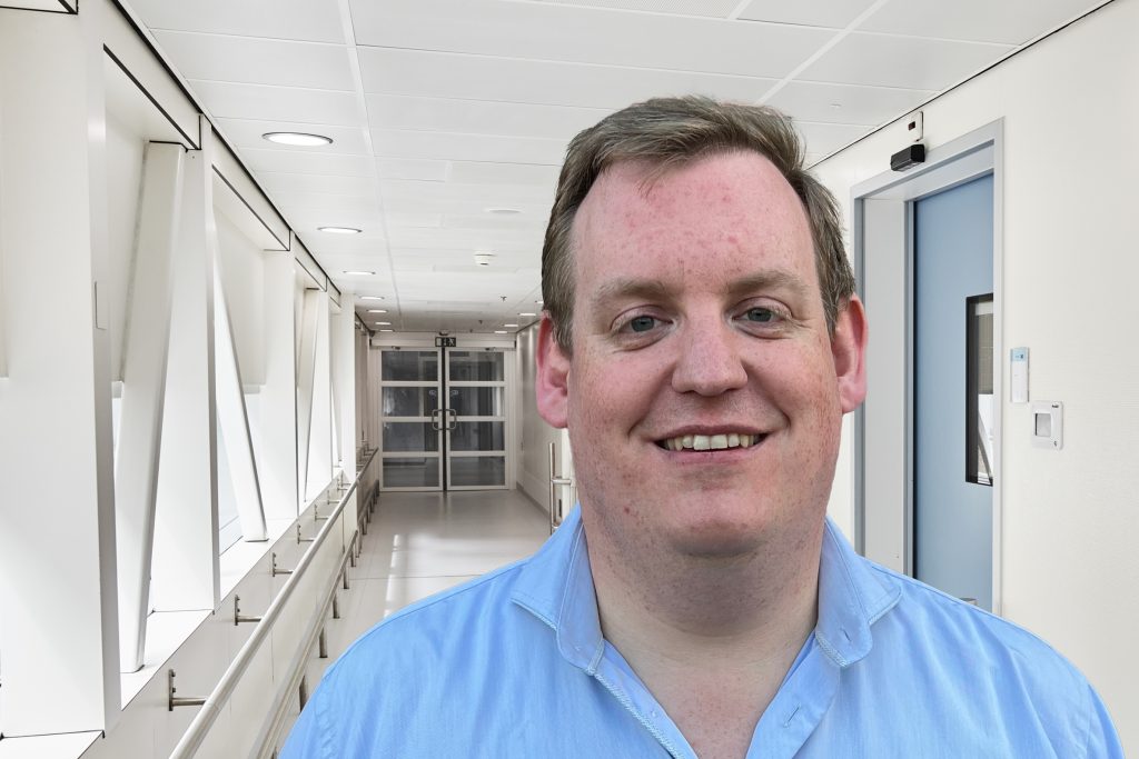 Man met kort bruin haar en lichtblauw overhemd lacht naar de camera in een lichte, moderne gang met veel natuurlijk licht.