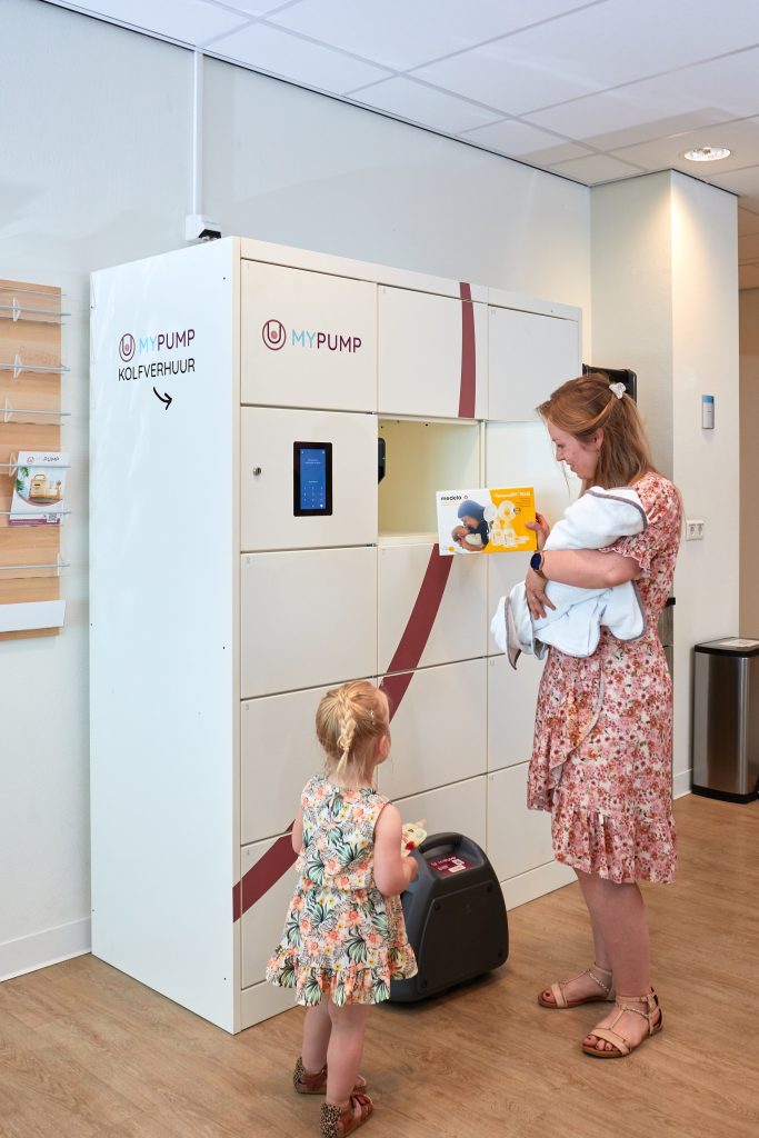Een vrouw met baby naast een meisje opent bij MyPump-locker een deur en bekijkt een gele handleiding; benodigdheden binnen handbereik.