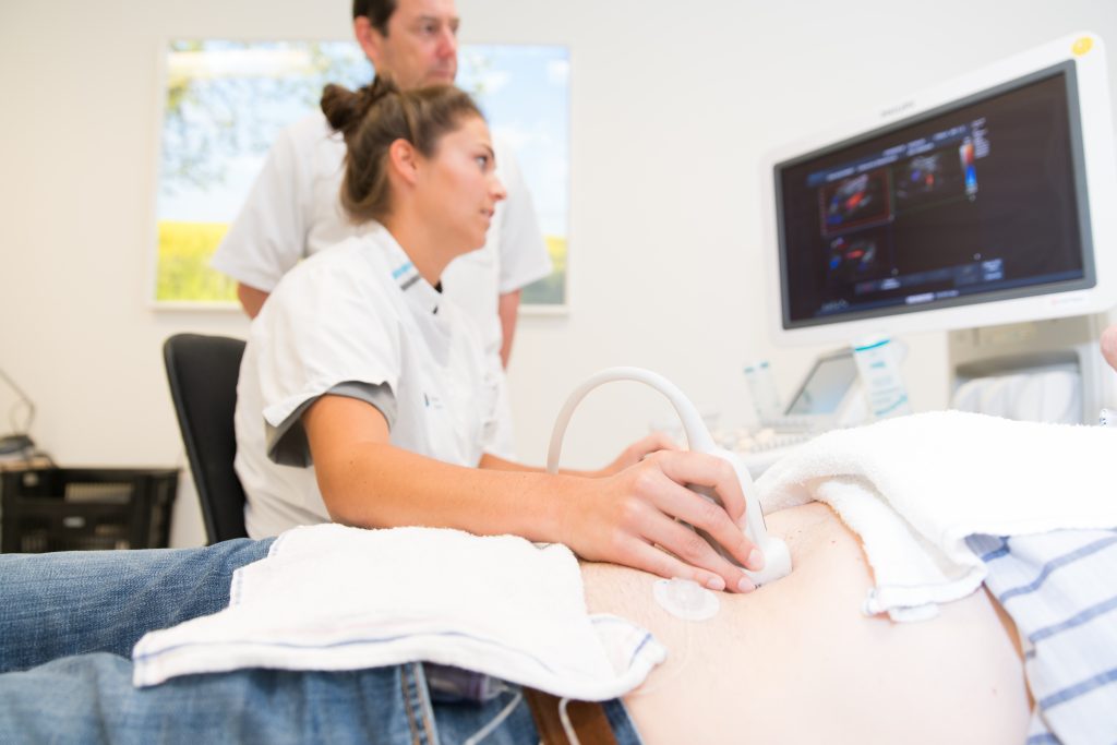 Arts voert buikecho uit bij patiënt op onderzoekstafel; collega kijkt toe. Computermodellen voorspellen risico op aneurysma.