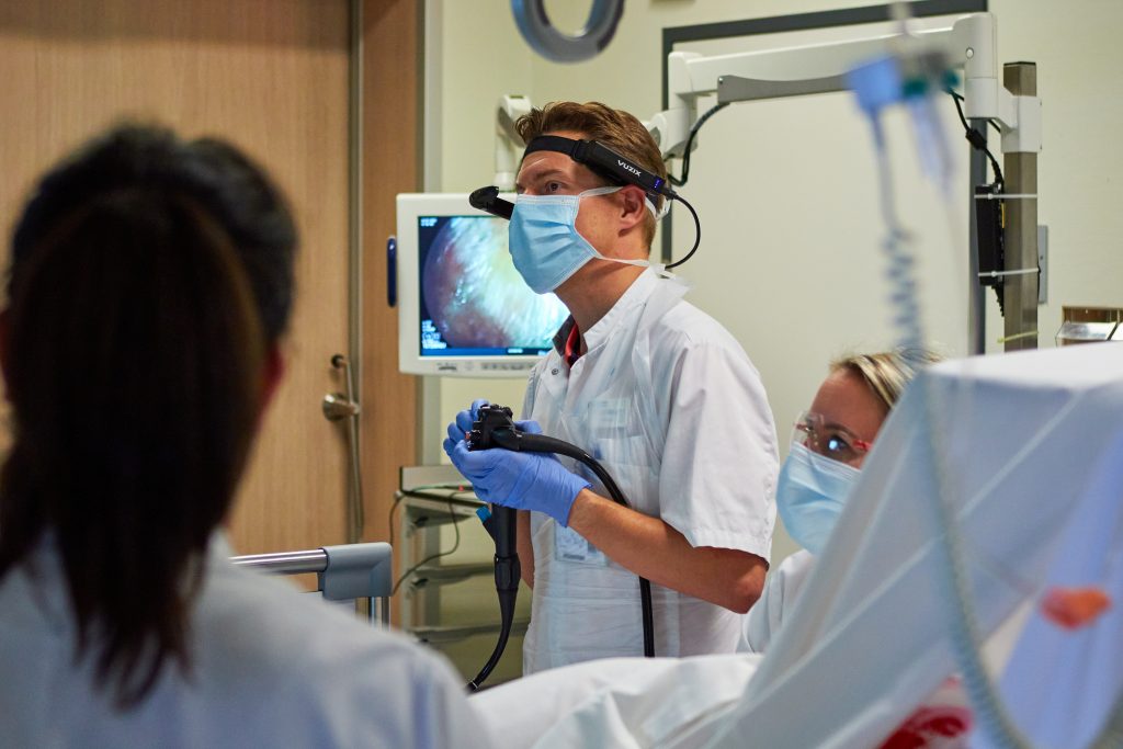 Drie specialisten met mondmaskers en handschoenen voeren een ingreep uit, met medische apparatuur en monitor in de operatiekamer.